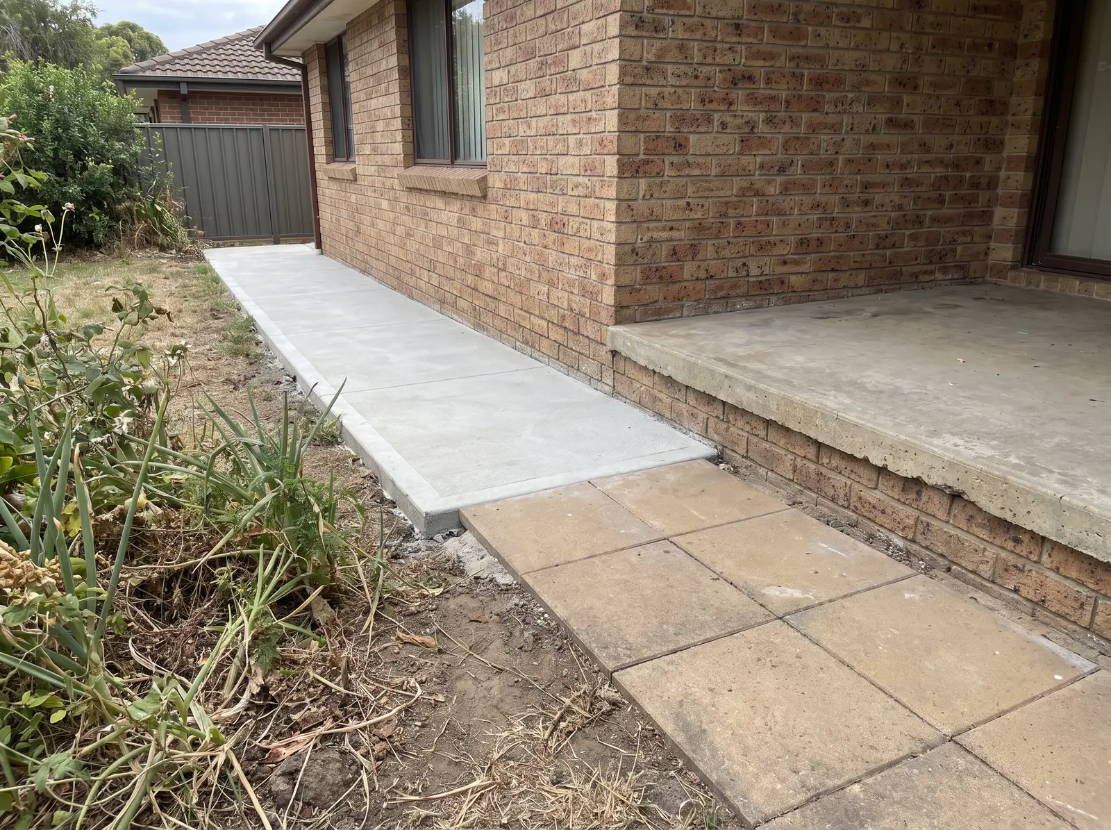 A concrete path and paving laid hard against a brick house wall, covering the slab edge